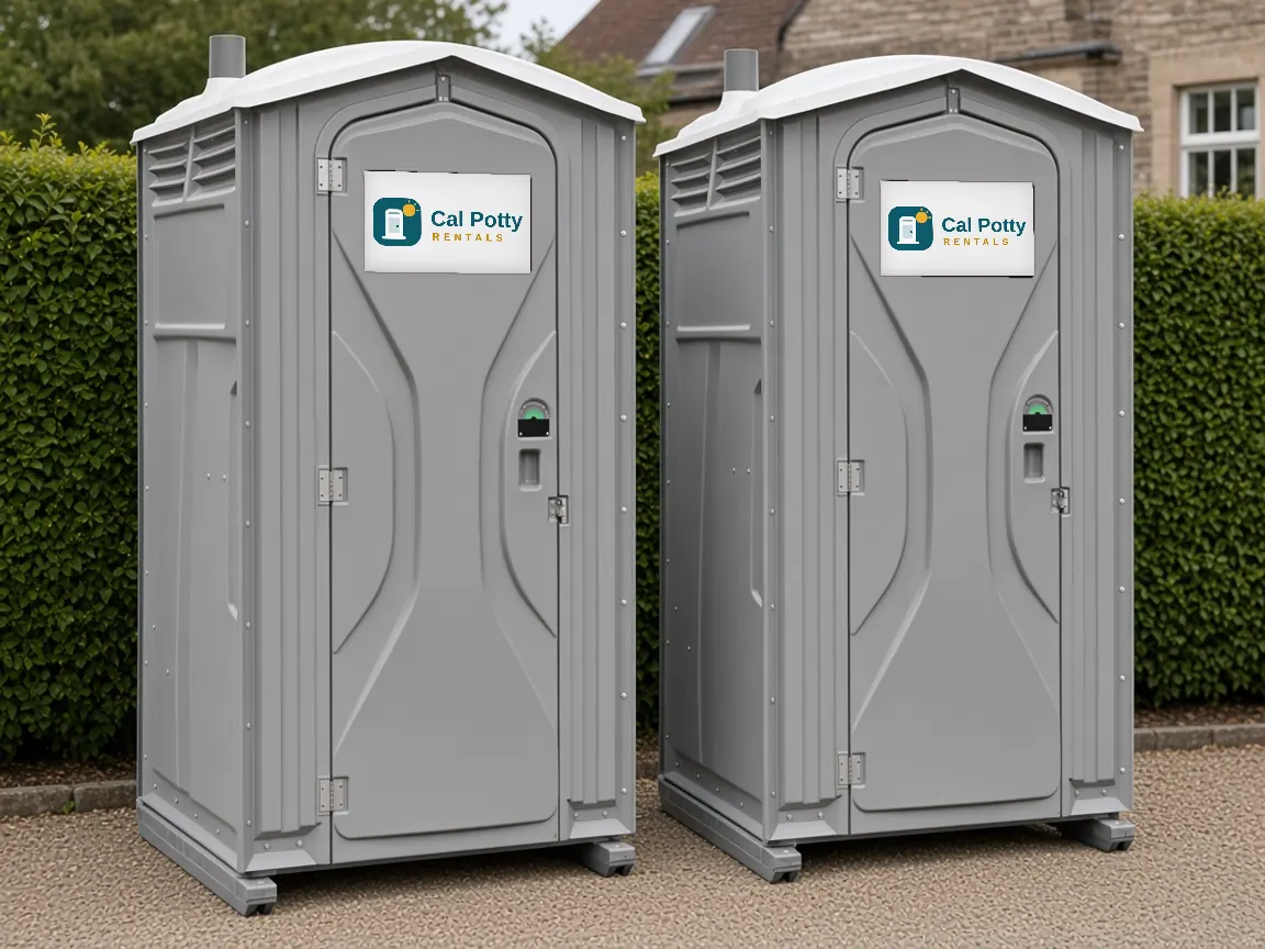 A pair of grey portable toilets on a gravel drive in front of a tall hedge at an estate
