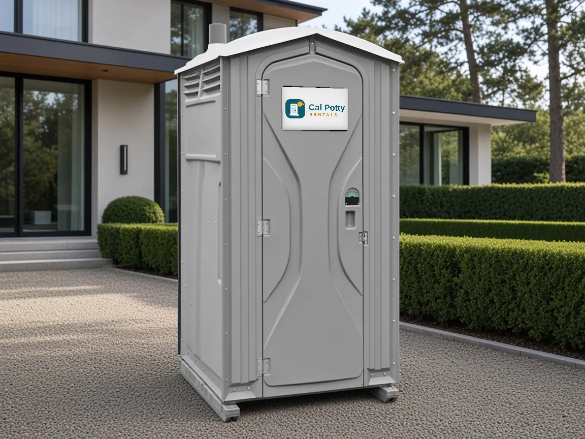A grey porta potty on a gravel drive in front of a modern home with hedges, an example setup for porta potty rental in Portola Valley