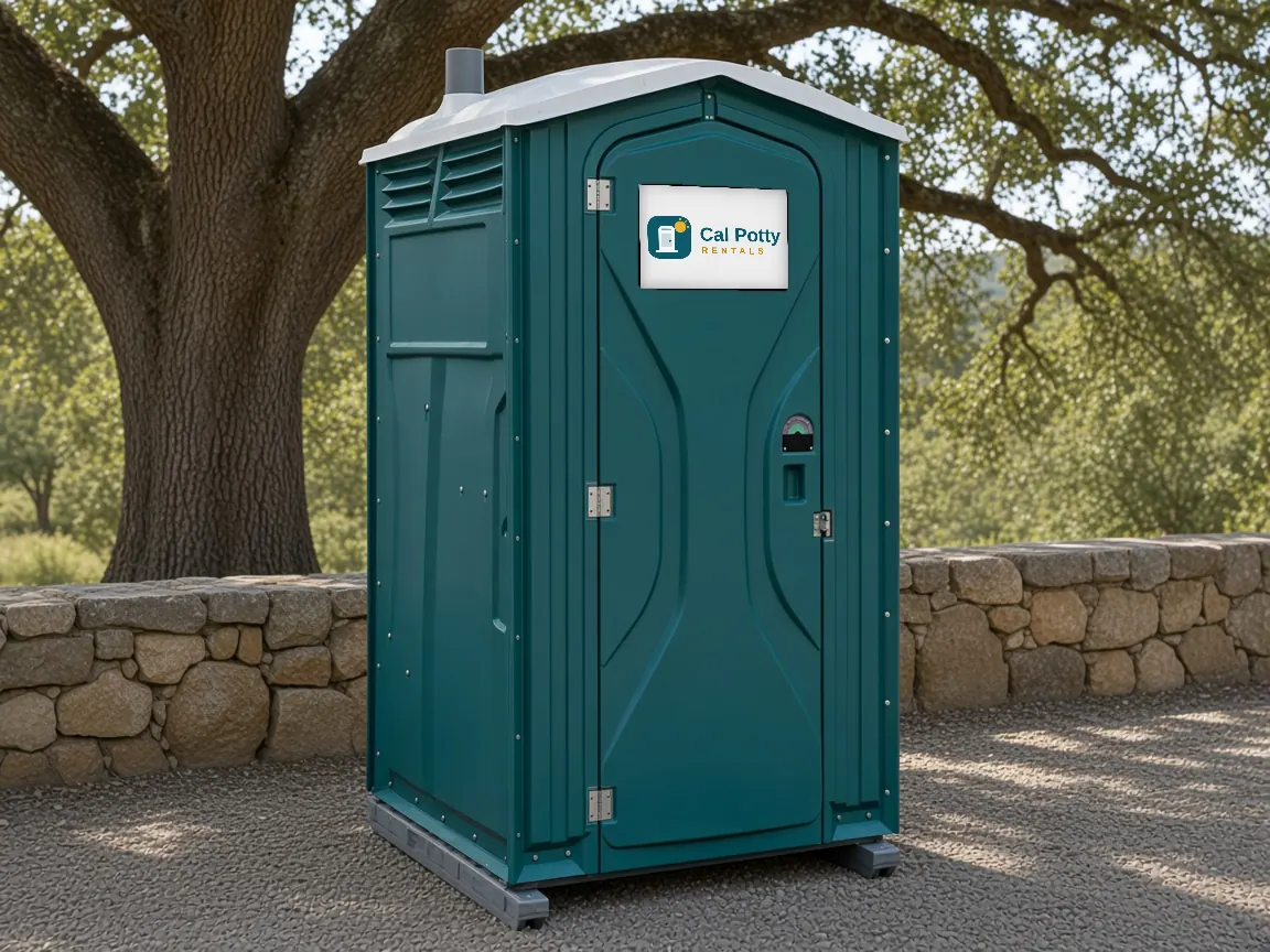 A teal porta potty beside a stone wall under a large oak tree